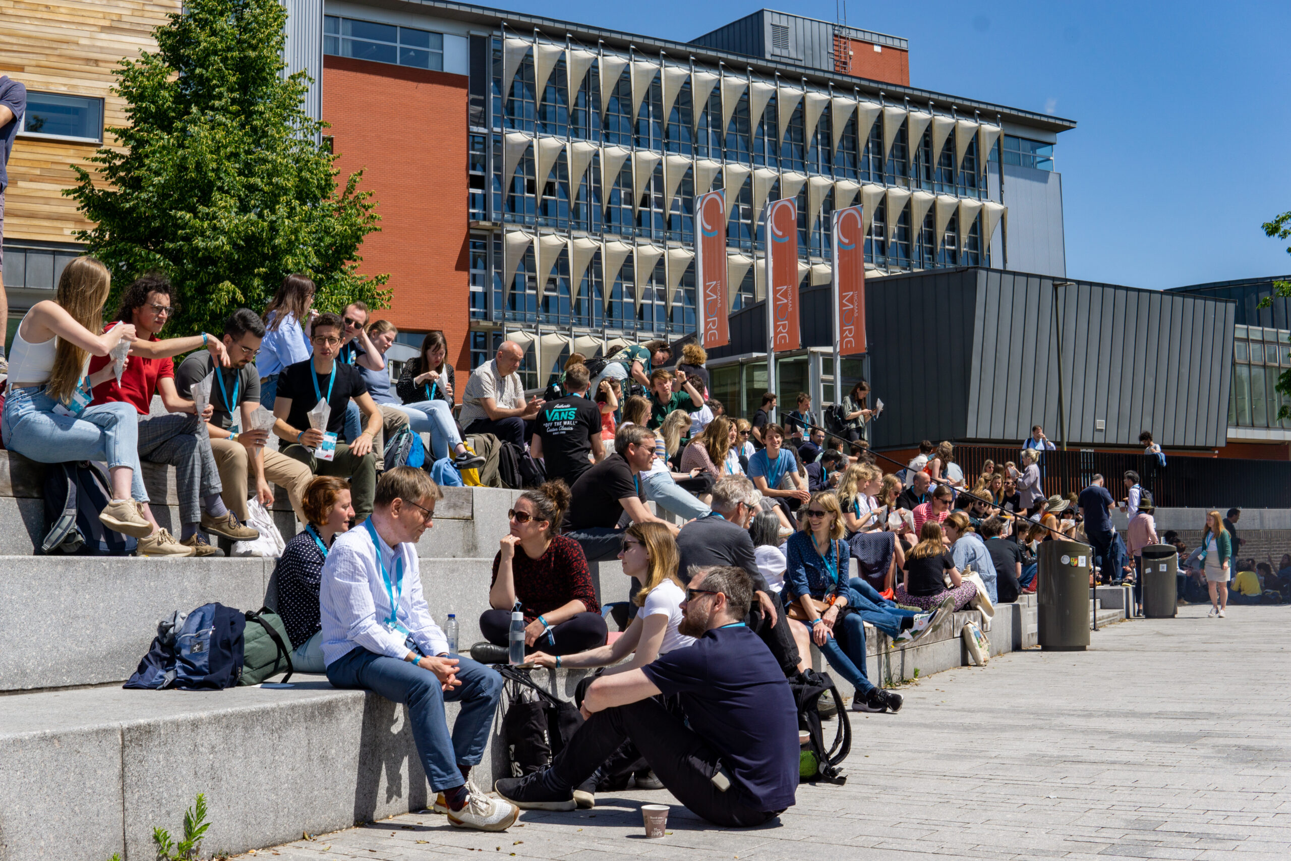 A large group of conference-goers, wearing light blue Dataharvest lanyards, eating lunch on the steps outside the conference venue.